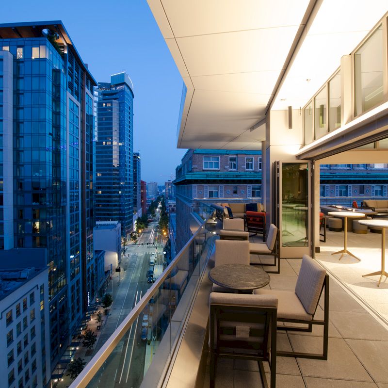 A modern high-rise cityscape viewed from a terrace at dusk, with tables and chairs, overlooking a street lined with buildings and lights.