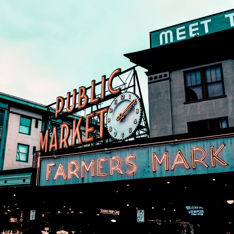 The image shows a neon sign for "Public Market" and "Farmers Market" with a clock on it, set against a building background.