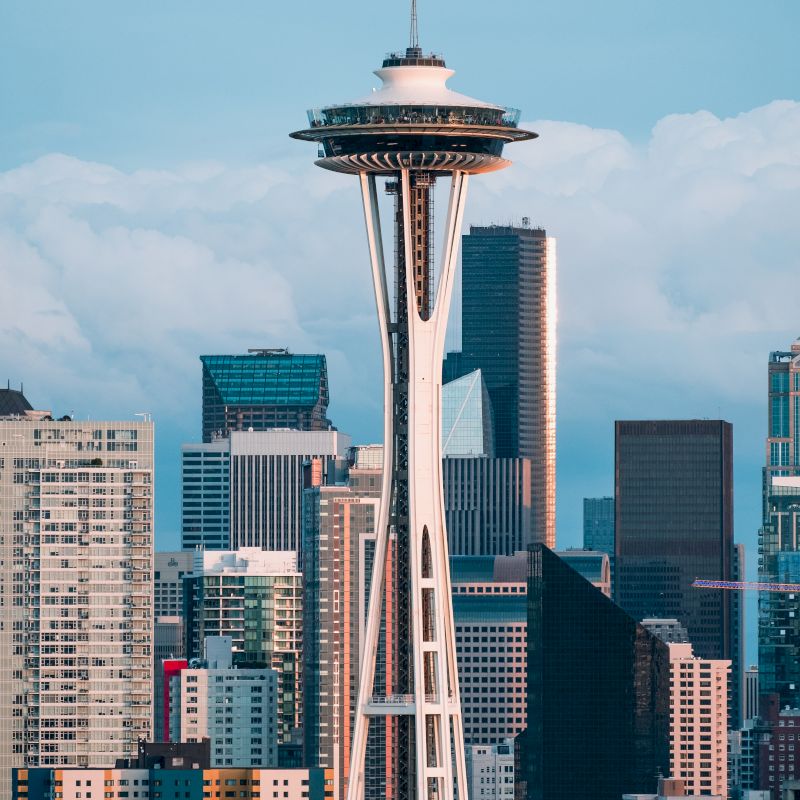 The image shows the Space Needle with a backdrop of skyscrapers, set against a sky with clouds. It is a prominent landmark in Seattle.