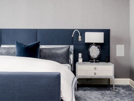 The image shows a modern bedroom with a blue and white color scheme, featuring a bed, pillows, nightstand, lamp, and carpeted floor.