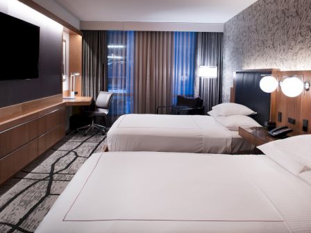 A modern hotel room features two beds, a wall-mounted TV, desk with chair, large window, and stylish decor in a neutral color palette.