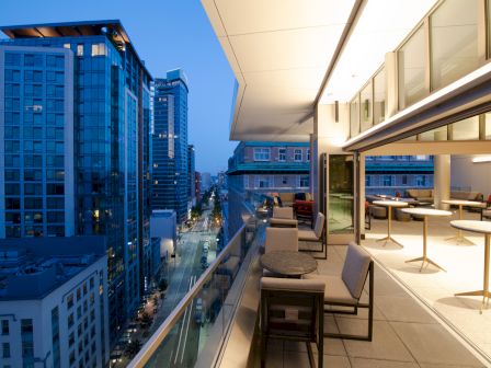 A rooftop terrace with seating overlooks a cityscape at dusk, featuring modern buildings and a view down a street.