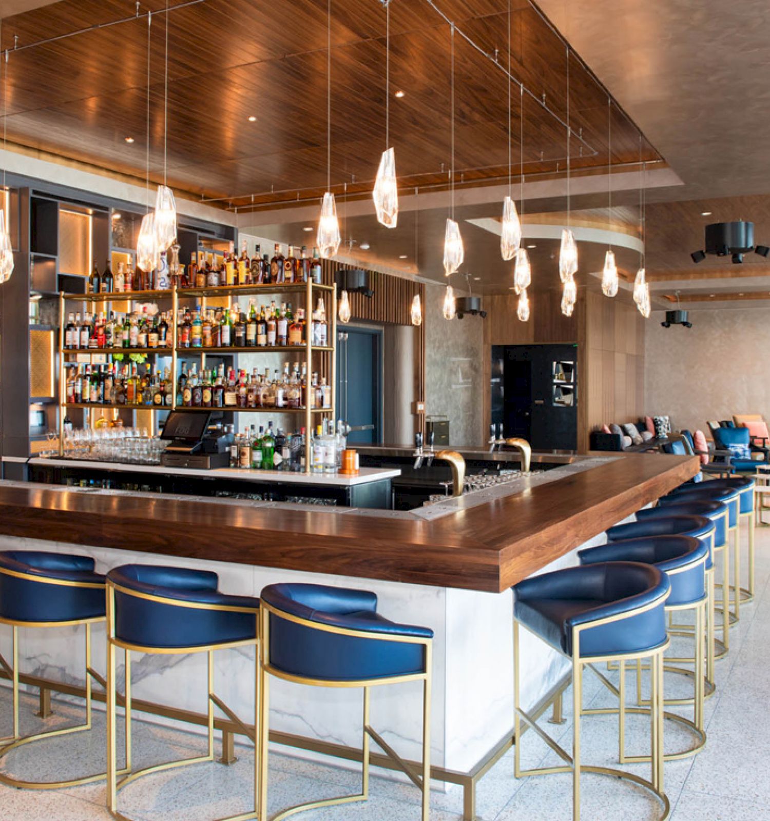 The image shows a modern bar with blue chairs, pendant lights, and a wide selection of bottles behind the counter in an inviting space.