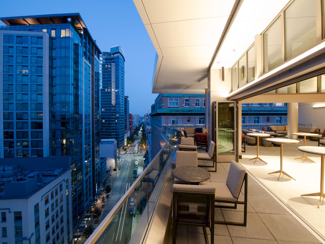 A modern balcony view in an urban setting, featuring outdoor seating and glass railings with city skyscrapers in the background.