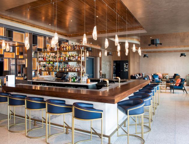 The image shows a modern bar with a central counter, blue bar stools, hanging lights, and a well-stocked liquor display, set in a stylish interior.