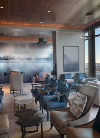 The image shows a modern lounge with elegant seating, large windows offering a scenic view, and a mural of a city skyline on the wall.