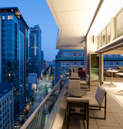A modern rooftop terrace with seating overlooks a cityscape with illuminated buildings at twilight.