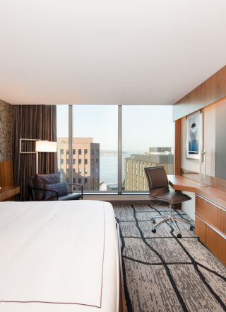 A modern hotel room with a large bed, desk, chair, and wall-mounted TV. The window offers a city view with buildings visible outside.