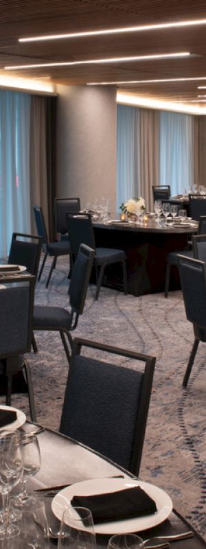 The image shows an elegant banquet hall with round tables set for a formal event, featuring dark chairs, dim lighting, and draped windows.