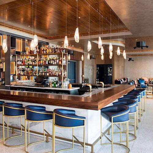 A modern bar with blue stools, hanging lights, and a well-stocked shelf. The setting is stylish and inviting, great for socializing.