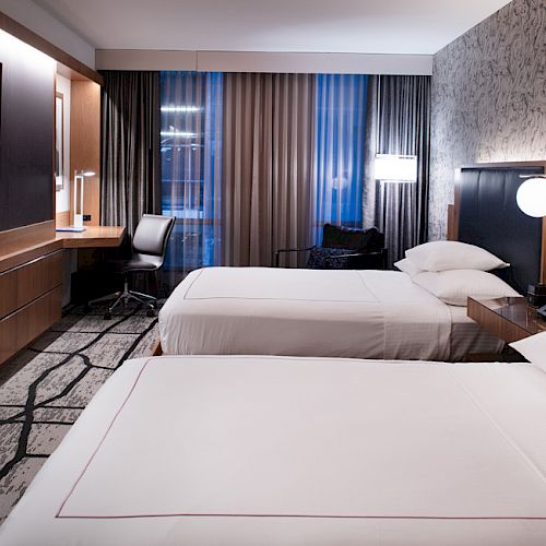 The image shows a modern hotel room with two beds, a TV, a desk, and stylish decor, featuring patterned wallpaper and curtains.