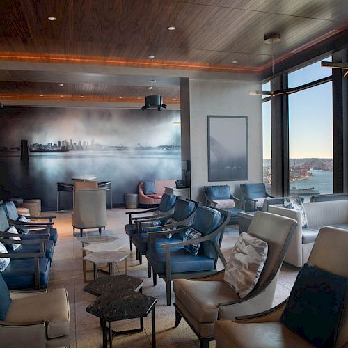 A stylish lounge with comfy seating, a cityscape mural, large windows, and a scenic view of the water and Ferris wheel outside.