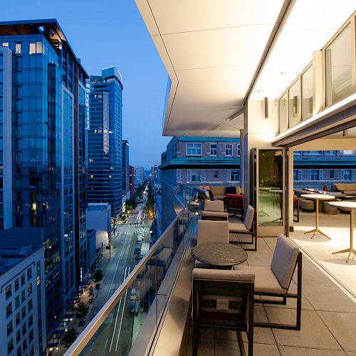 The image shows a modern balcony with seating and tables, overlooking a cityscape with tall buildings and a street below at dusk.