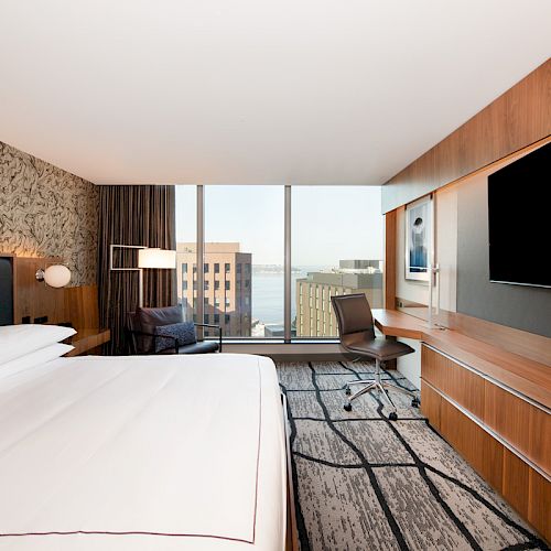 A modern hotel room with a large bed, a desk and chair, a wall-mounted TV, and a window with a view of city buildings and water.