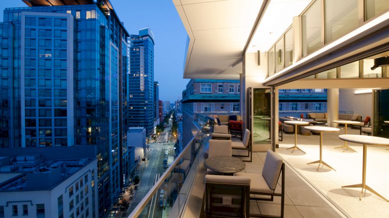 A modern balcony overlooking a cityscape at dusk, featuring outdoor seating and tables, with illuminated buildings lining the street below.