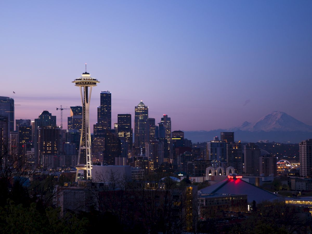 A city skyline at dusk with a prominent tower and a mountain in the background, under a purple and blue sky.