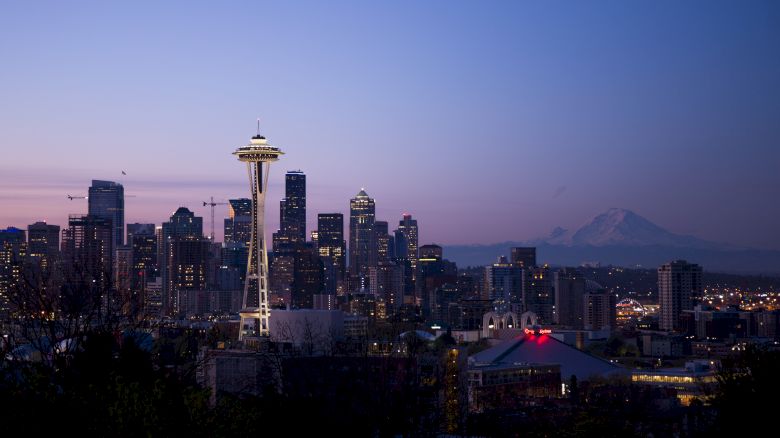 A city skyline at dusk with a prominent tower and a mountain in the background, under a purple and blue sky.