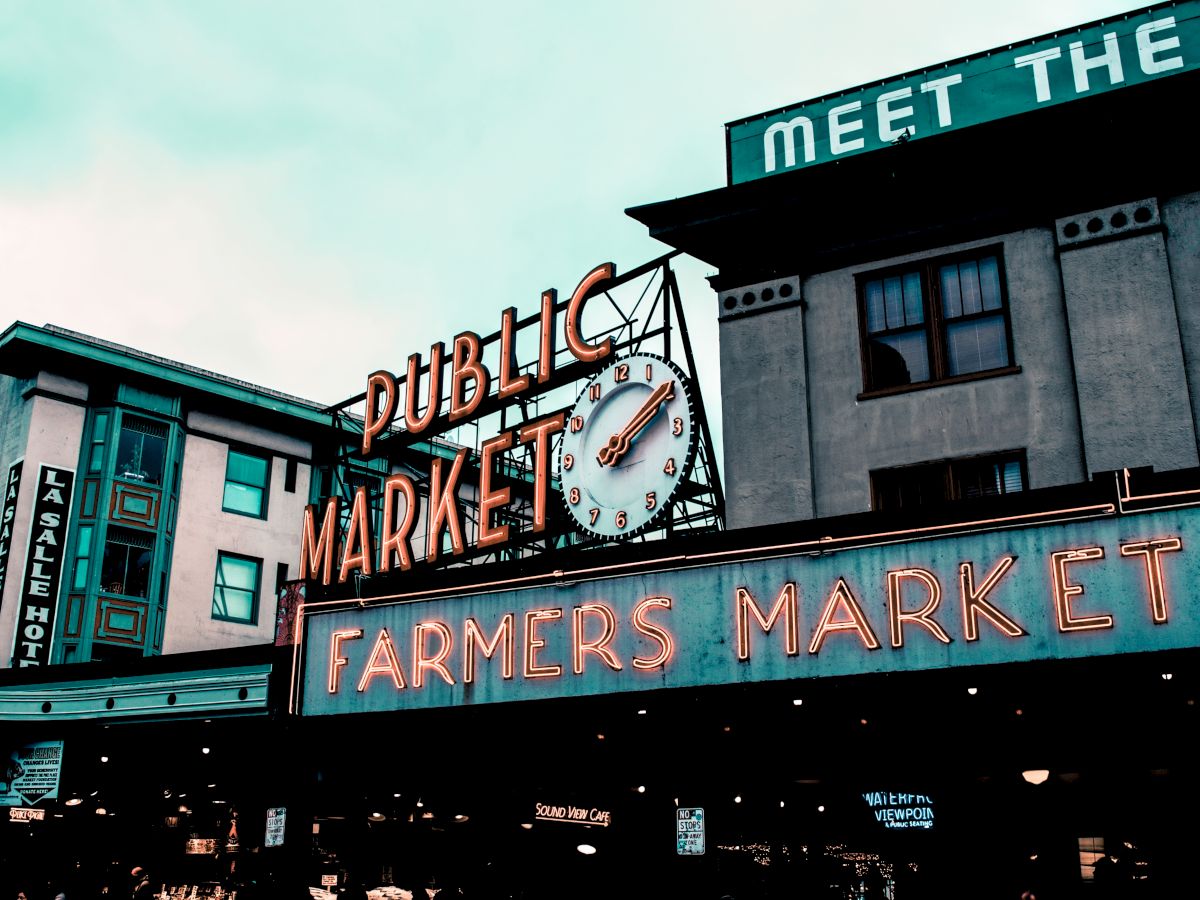 The image shows an iconic sign for a public farmers market with a large clock, set against a backdrop of buildings.
