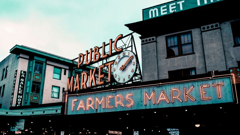 The image shows an iconic sign for a public farmers market with a large clock, set against a backdrop of buildings.