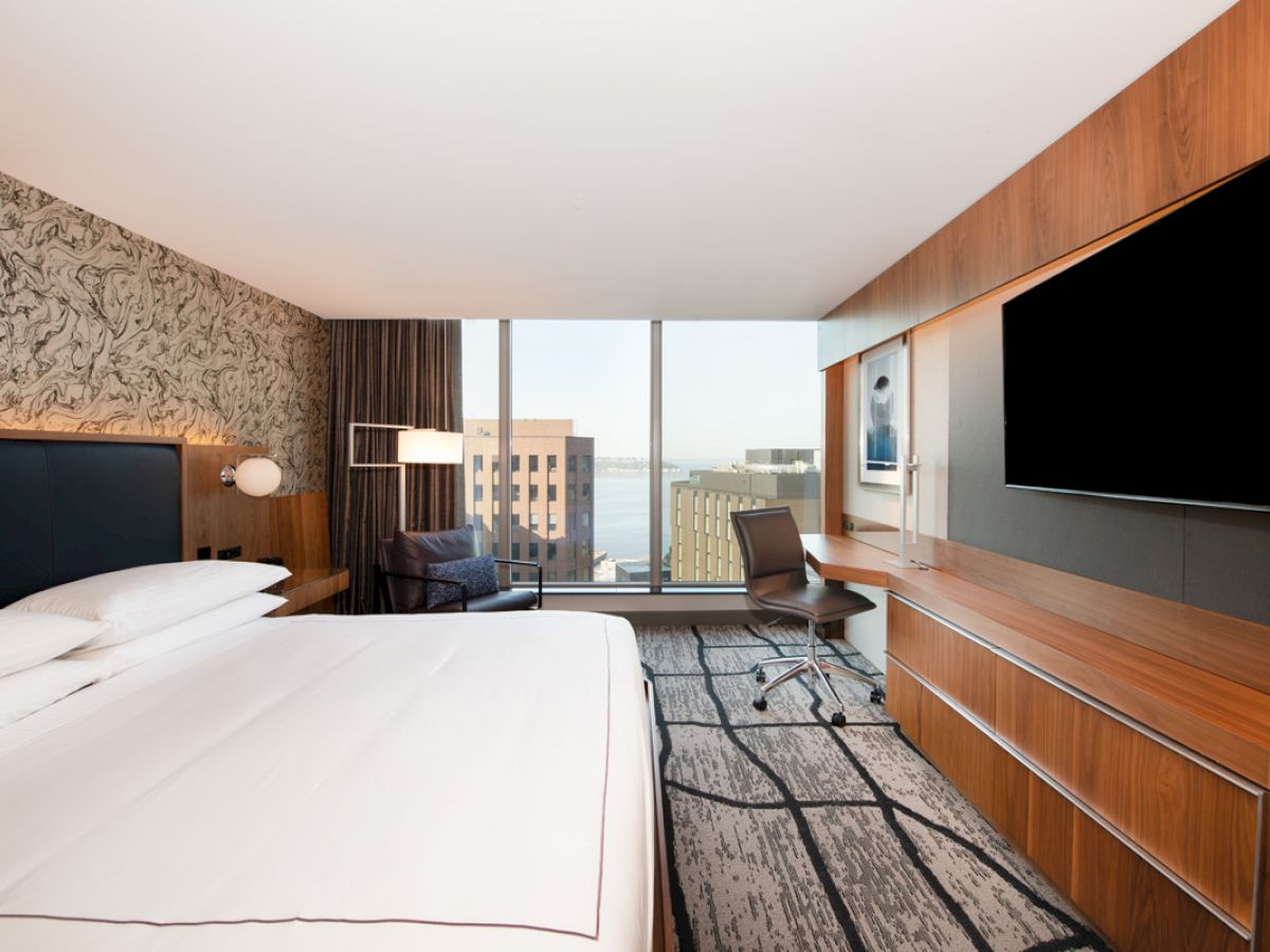A modern hotel room with a large bed, TV, desk, and window overlooking city buildings and water.