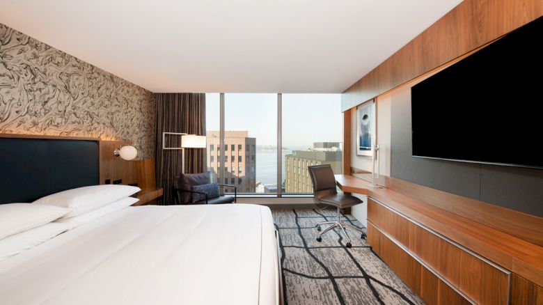 A modern hotel room with a large bed, TV, desk, and window overlooking city buildings and water.