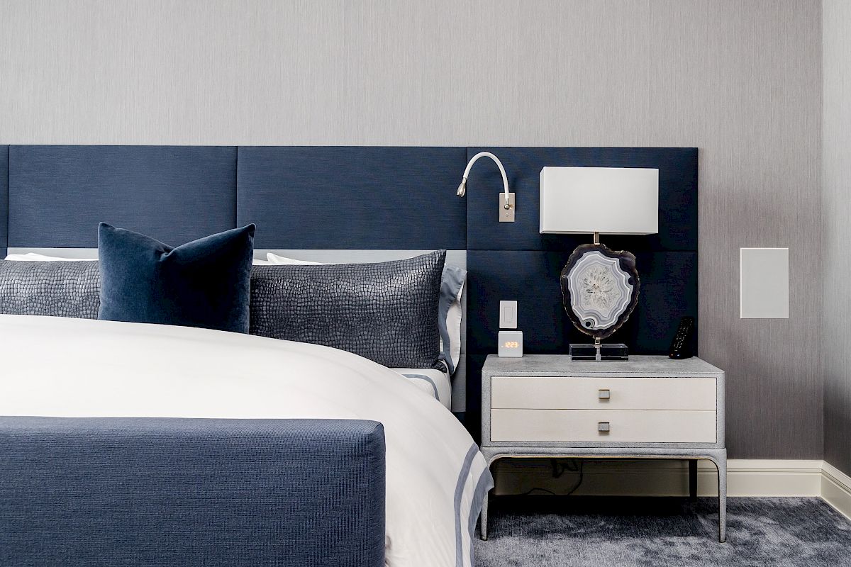 A modern bedroom with a blue upholstered headboard, white bedding, gray carpet, and a nightstand with a lamp and decorative objects.