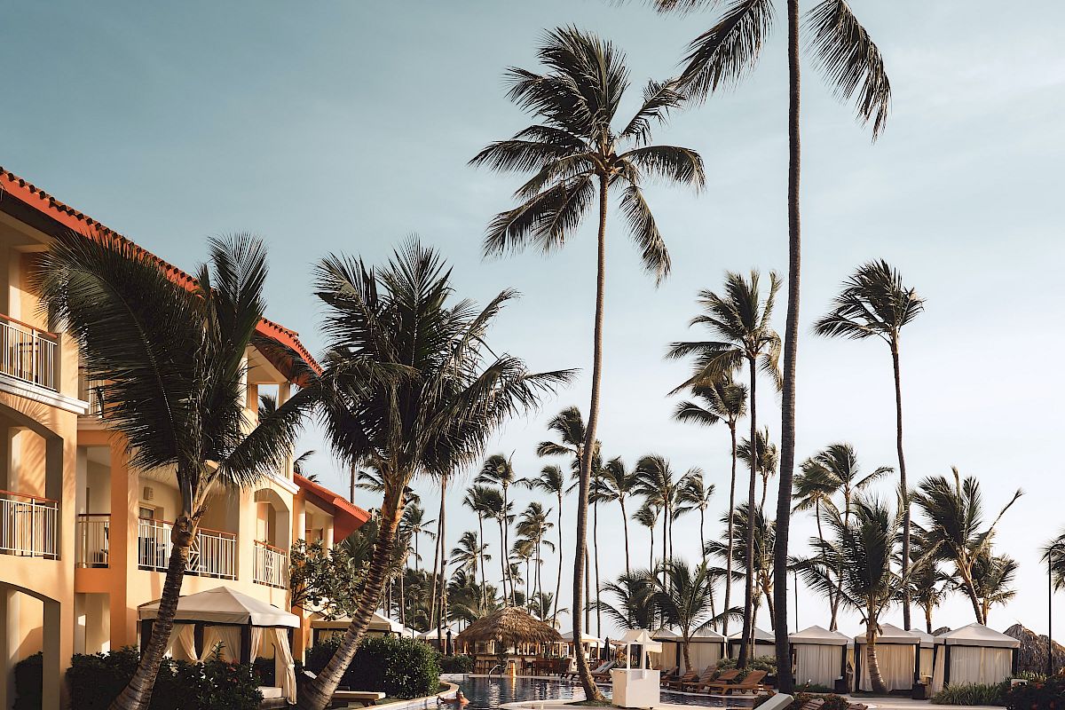 A tropical resort pool scene with lounge chairs, palm trees, and a building under a clear sky.