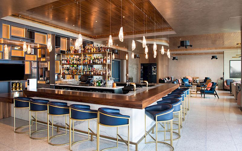 A modern bar with a sleek wooden counter, blue barstools, decorative lighting, shelves stocked with bottles, and a seating area in the back.