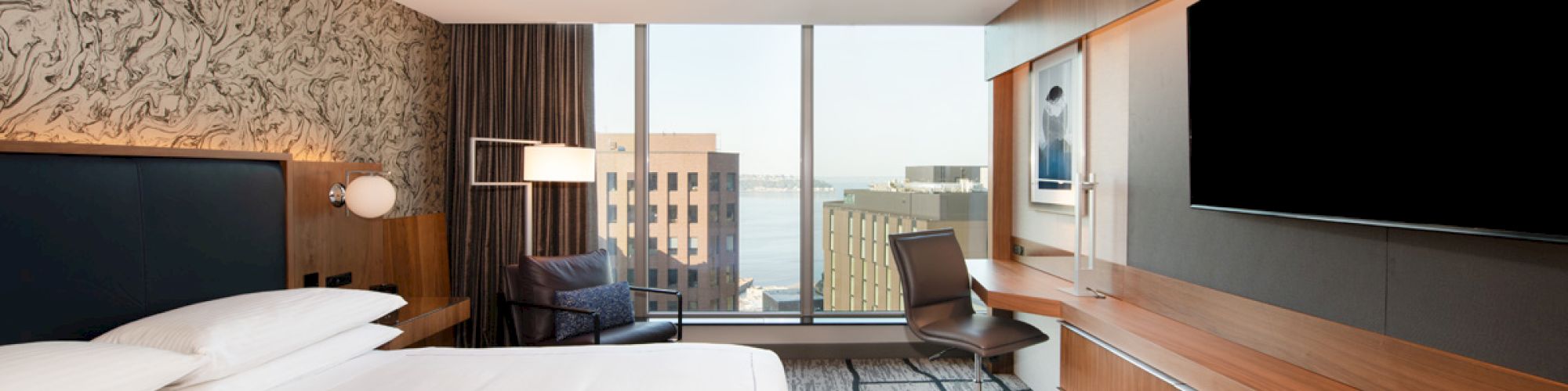 A modern hotel room with a large bed, wall-mounted TV, desk, chair, and a window with a city view in the background.